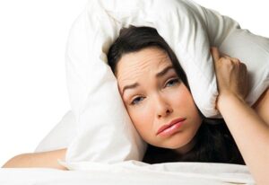 Woman lying in bed covering her ears with a white pillow, appearing frustrated by noise disturbance.