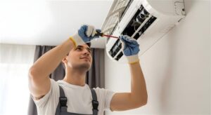 Technician in work uniform and protective gloves servicing an indoor air conditioning unit mounted on ceiling.