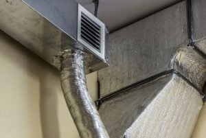 Flexible metal ductwork connected to ceiling-mounted air vent with insulation wrap visible on basement or utility room walls.