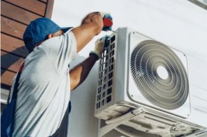 Technician in blue cap installing exterior air conditioning unit on residential wall.