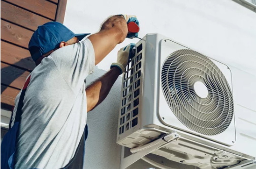 Technician in blue cap installing exterior air conditioning unit on residential wall.