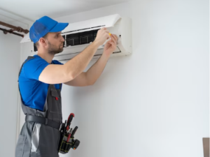 Skilled technician in blue uniform and cap installing or servicing wall-mounted air conditioning unit indoors.