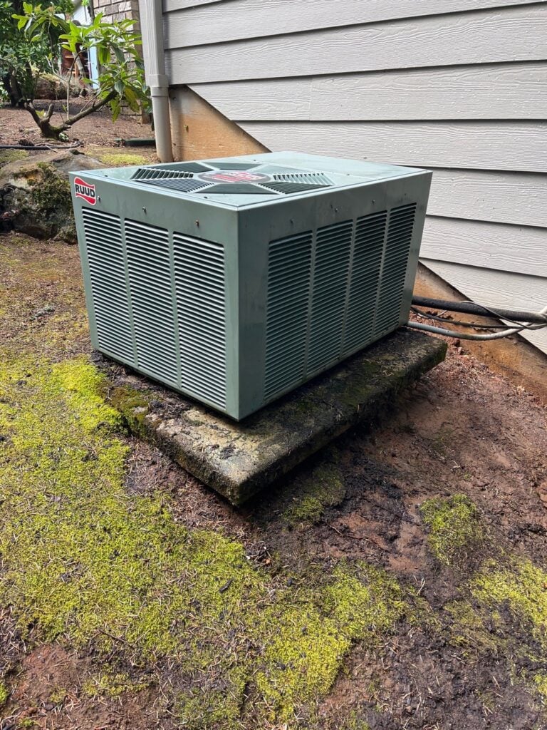 Ruud air conditioning condenser unit installed on concrete pad against house siding with moss growth.