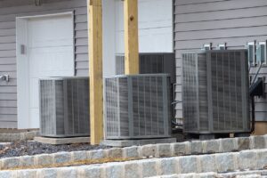 Multiple gray HVAC air conditioning units mounted on concrete foundation against residential siding with wooden support posts.