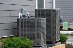 Two gray metal air conditioning condenser units installed against a house with gray siding and green landscaping shrubs.