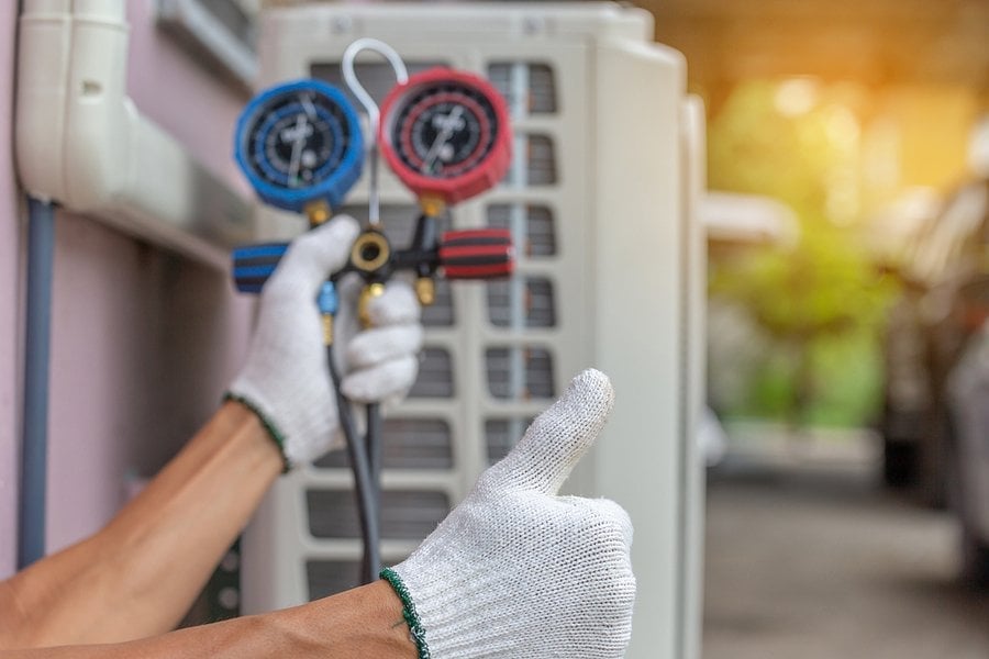 Technician in white gloves holding blue and red manifold gauges connected to an air conditioning unit for refrigerant measurement and diagnosis.