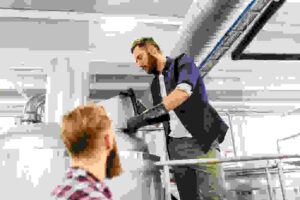 Two workers inspect equipment at a craft brewery facility, one standing on a ladder wearing safety gear while another observes from below.