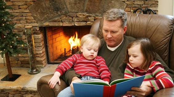 Adult and two children reading a blue book together by a warm fireplace during holiday season.