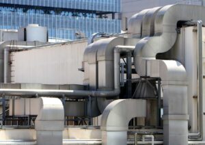 Industrial HVAC ductwork and ventilation pipes on rooftop with modern office building in background.
