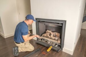 Technician in blue shirt and cap kneeling beside an open gas fireplace insert, adjusting internal components with tools during installation or maintenance.