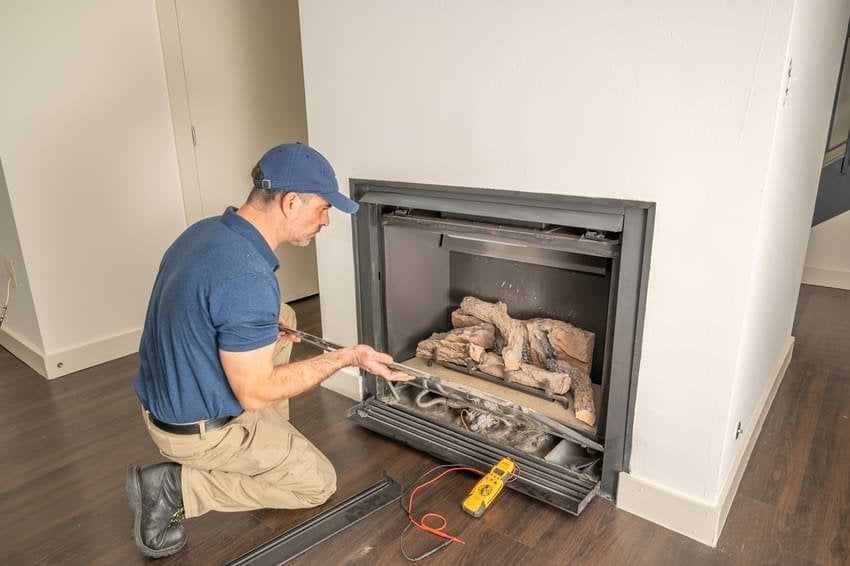 Technician in blue shirt and cap kneeling beside an open gas fireplace insert, adjusting internal components with tools during installation or maintenance.