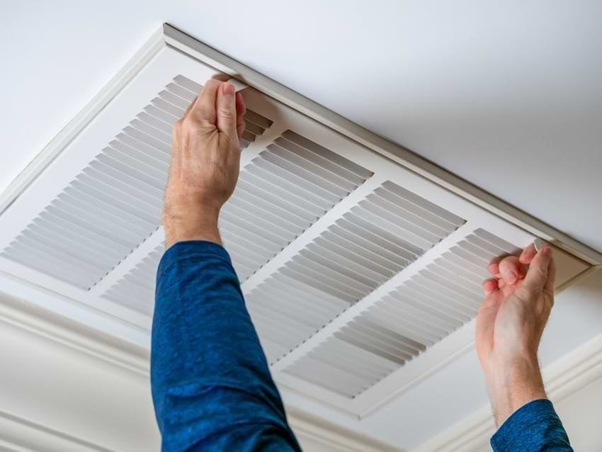 Person in blue shirt cleaning a ceiling air duct filter with both hands raised upward.