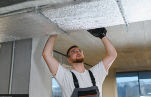 Worker installing insulation on ceiling pipes during commercial HVAC maintenance work indoors.
